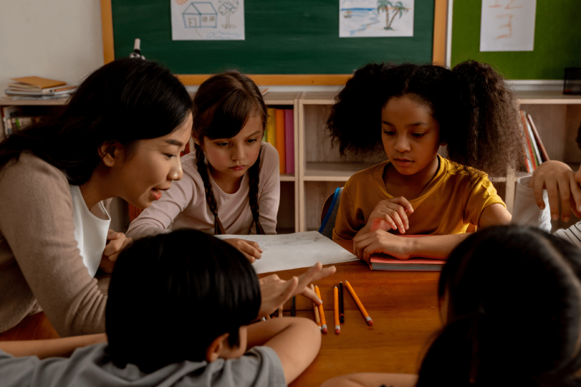 A diverse group of students are around a table, with a teacher counting pencils.