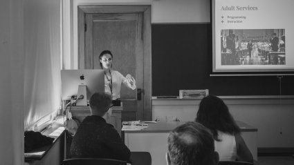  A student presenting at LEAP 2018 stands in front of a slide that reads "Adult Services."