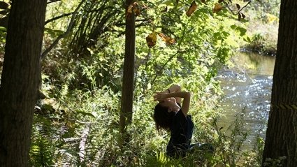 A student wearing dark clothing sits in the shadow of the woods next to a body of water, with head extended back and hands together at the forehead, and sun shining just beyond, 2018.