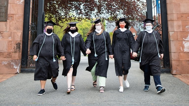 Mount Holyoke students in graduation garb and masks, striding through the gates with their hands locked, prepared to meet the world.