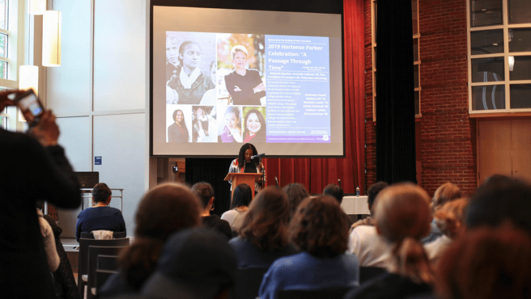 Kijua Sanders-McMurtry on stage speaking to the audience with a screen behind her and images from other Hortense Parker celebrations.