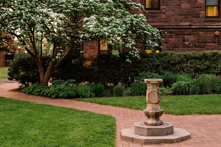 Sundial on campus with paths around it, trees, greens, and a brick building