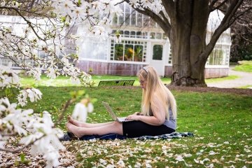 Student sitting on grass under a tree reading a book.